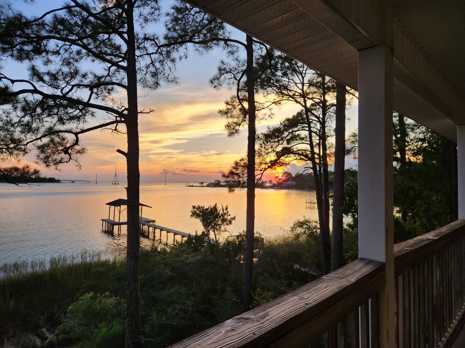 Sunset over East Bay seen from a covered balcony rail, framed by tall pines, with marsh grass below.