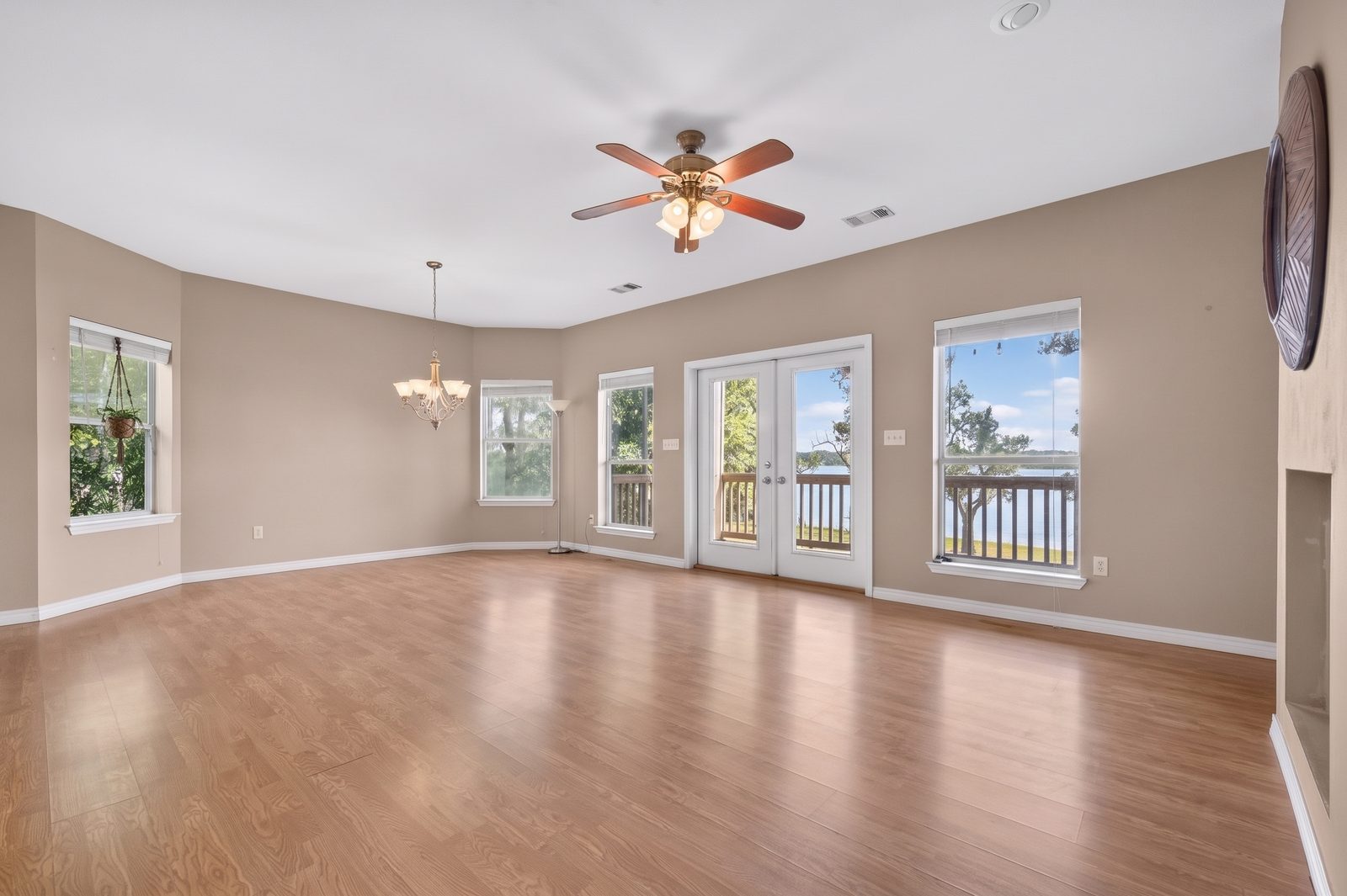 An empty living room with laminate floors and a ceiling fan, a dining area with a chandelier at left, and French doors and windows on the far wall looking out to a deck rail and the bay.