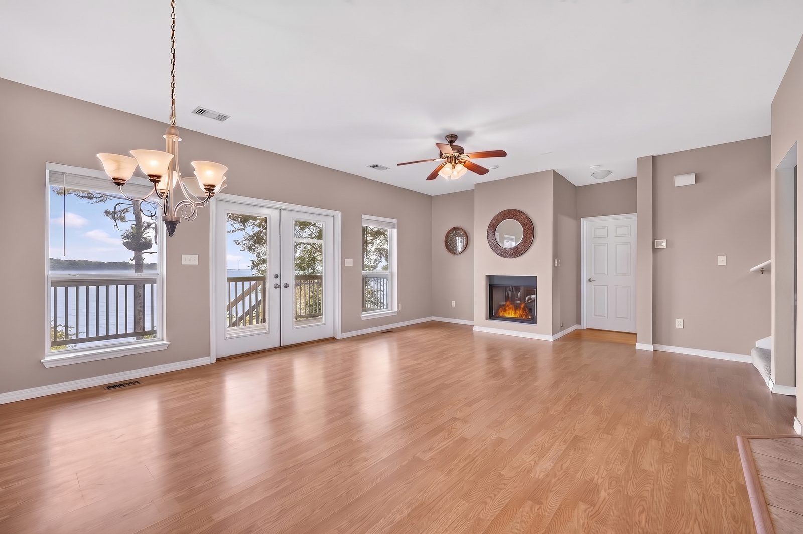 An empty living and dining area with laminate floors: a chandelier over the dining space, a window and French doors to a deck and the bay on the left, and a lit fireplace on the far wall.