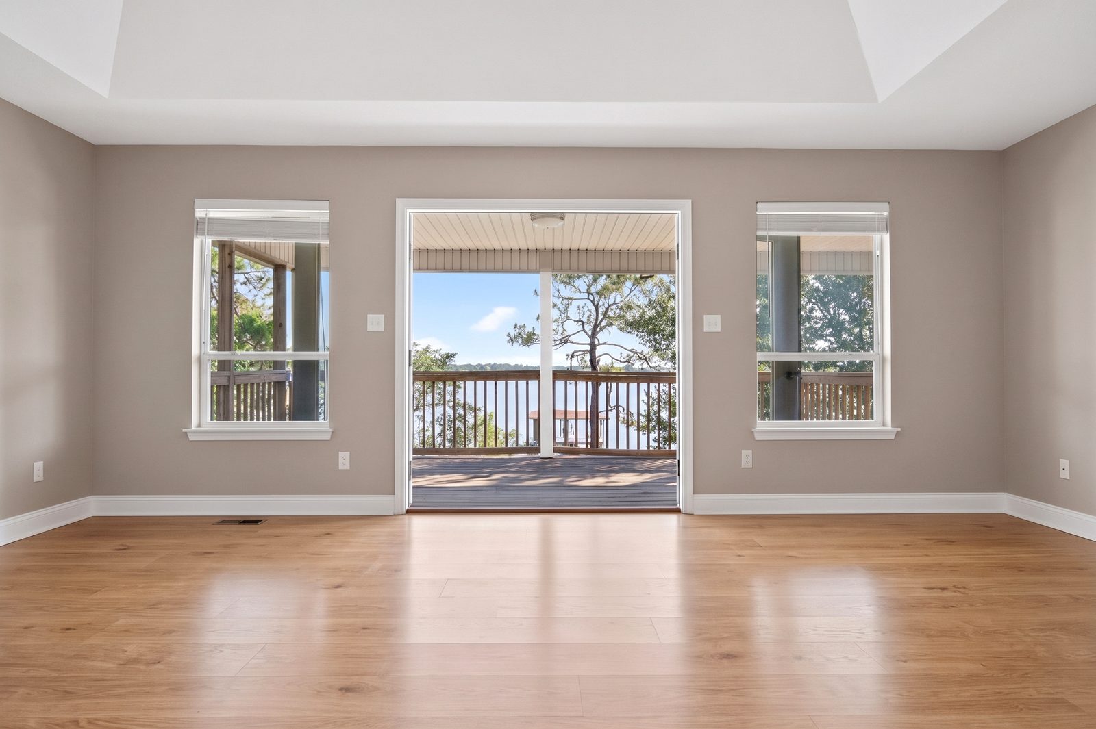 An empty room with a tray ceiling and wood-look floors, where a wide doorway between two windows opens onto a covered balcony overlooking the bay.