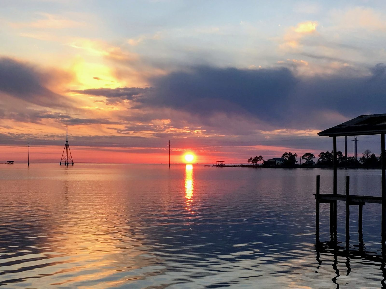 The sun sitting on the horizon over calm open water, its reflection running toward the camera, with towers standing in the water and the roof and pilings of a covered dock in silhouette at right.