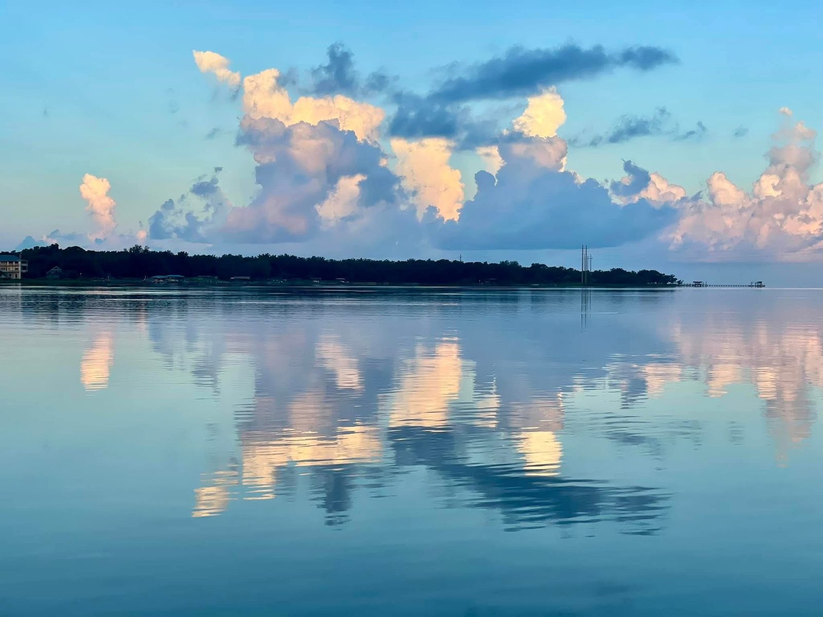 Tall white and pink clouds mirrored in flat, calm water, with a low wooded shoreline and a long pier on the far side.