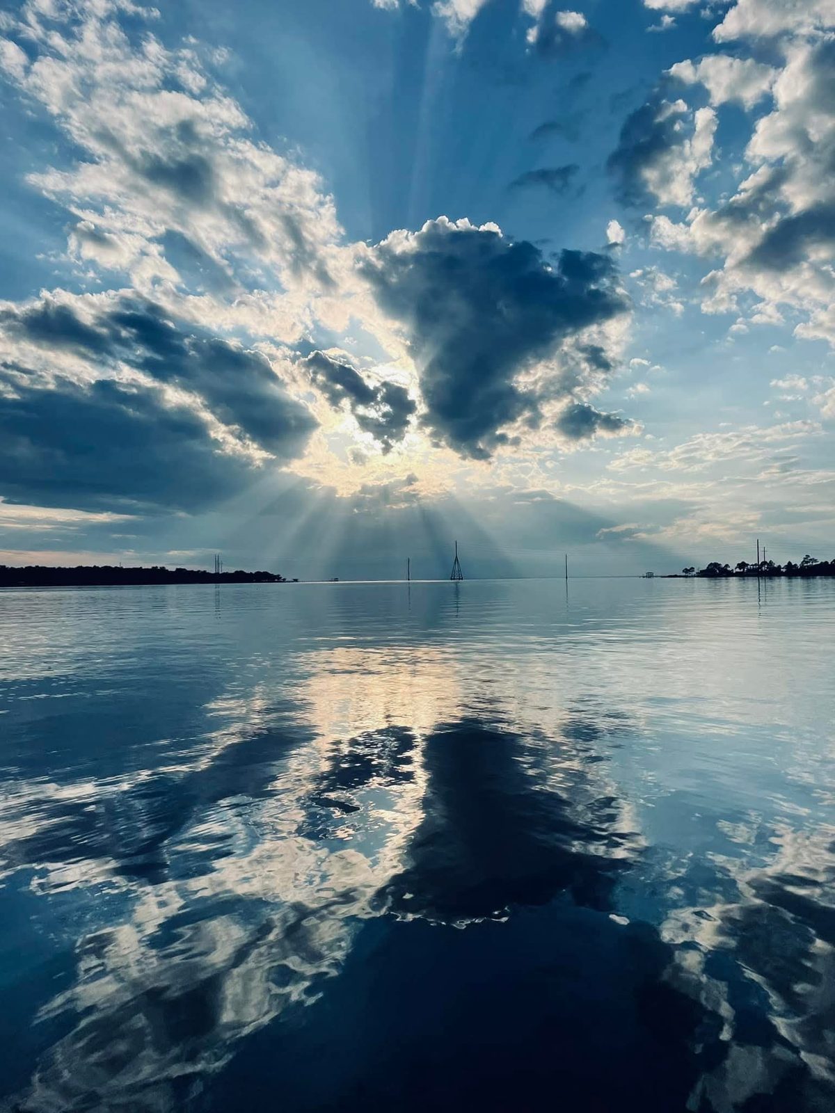 Sunbeams fanning out from behind a dark cloud over open water, the clouds mirrored in the calm surface, with towers on the horizon.