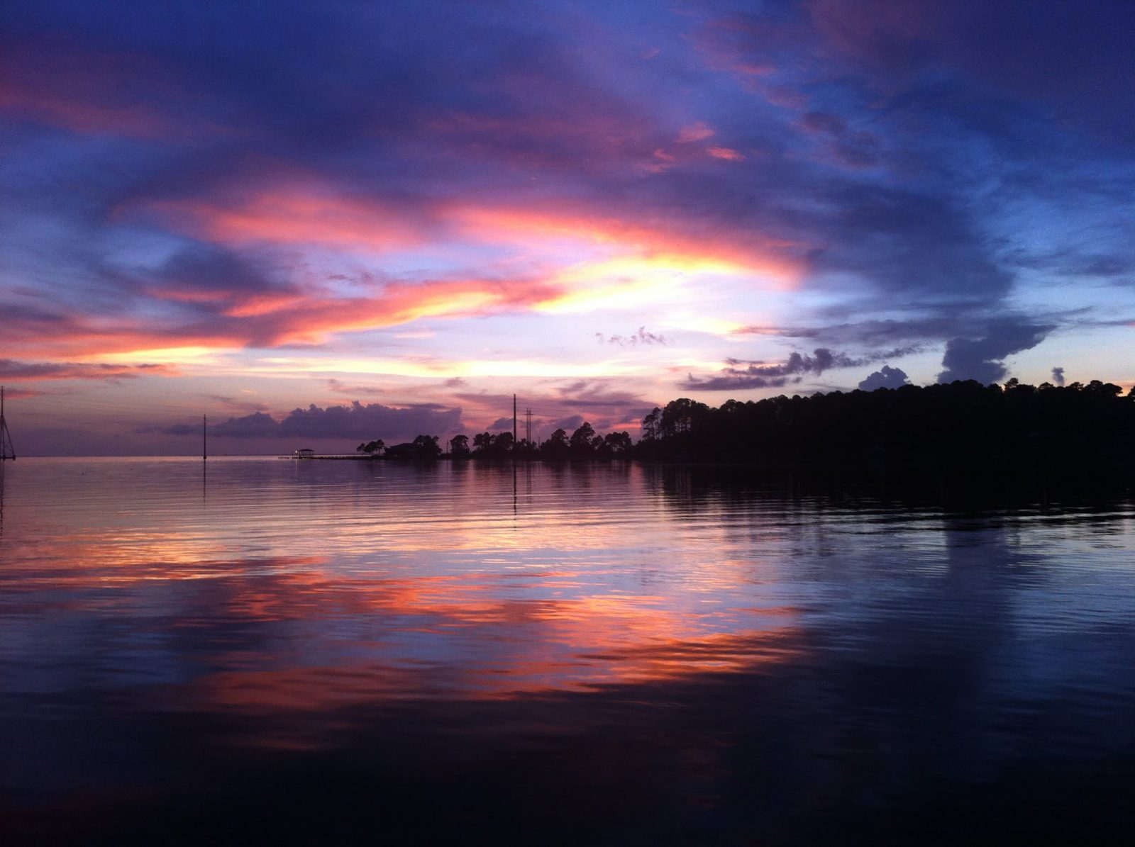 Pink and violet sunset clouds reflected in calm water, with a tree-lined point in silhouette at right.
