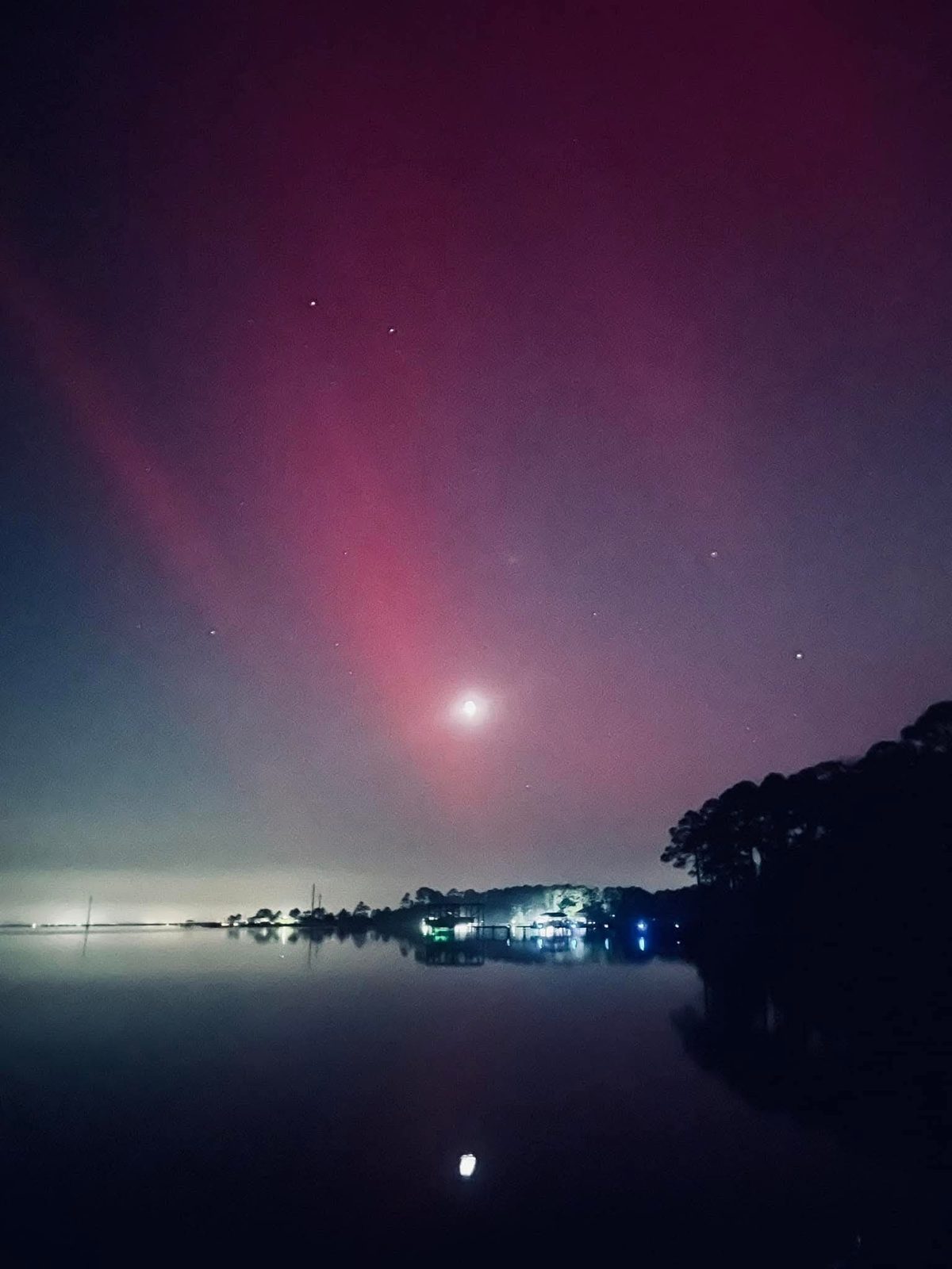 A night sky glowing pink and violet over still water, with a few stars, the lights of docks along the shore, and dark trees at right.