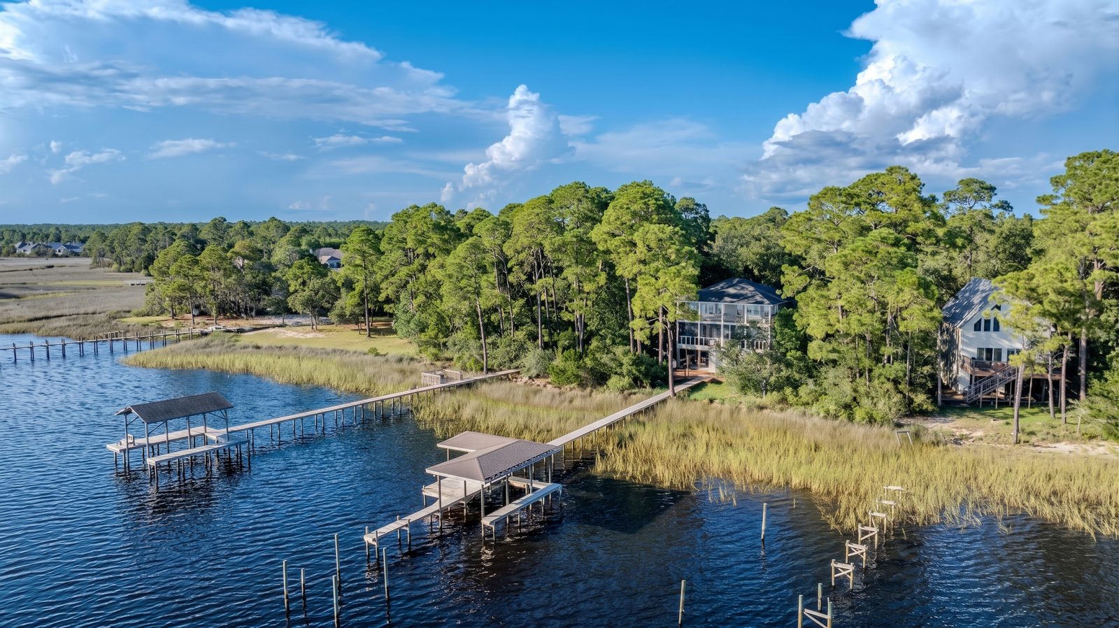 Aerial from over East Bay: the covered dock and boat slip in the foreground, a boardwalk crossing the marsh grass to a three-story house in tall pines, with a neighboring house at right.