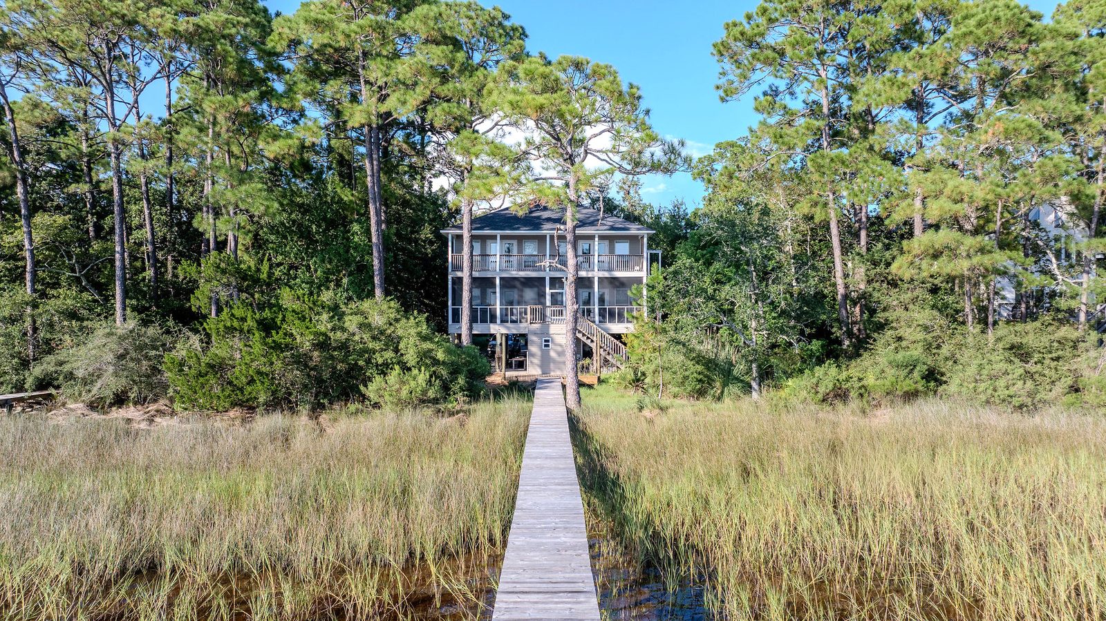 A long wooden boardwalk running straight through tall marsh grass to the back of a three-story house on pilings among tall pines.