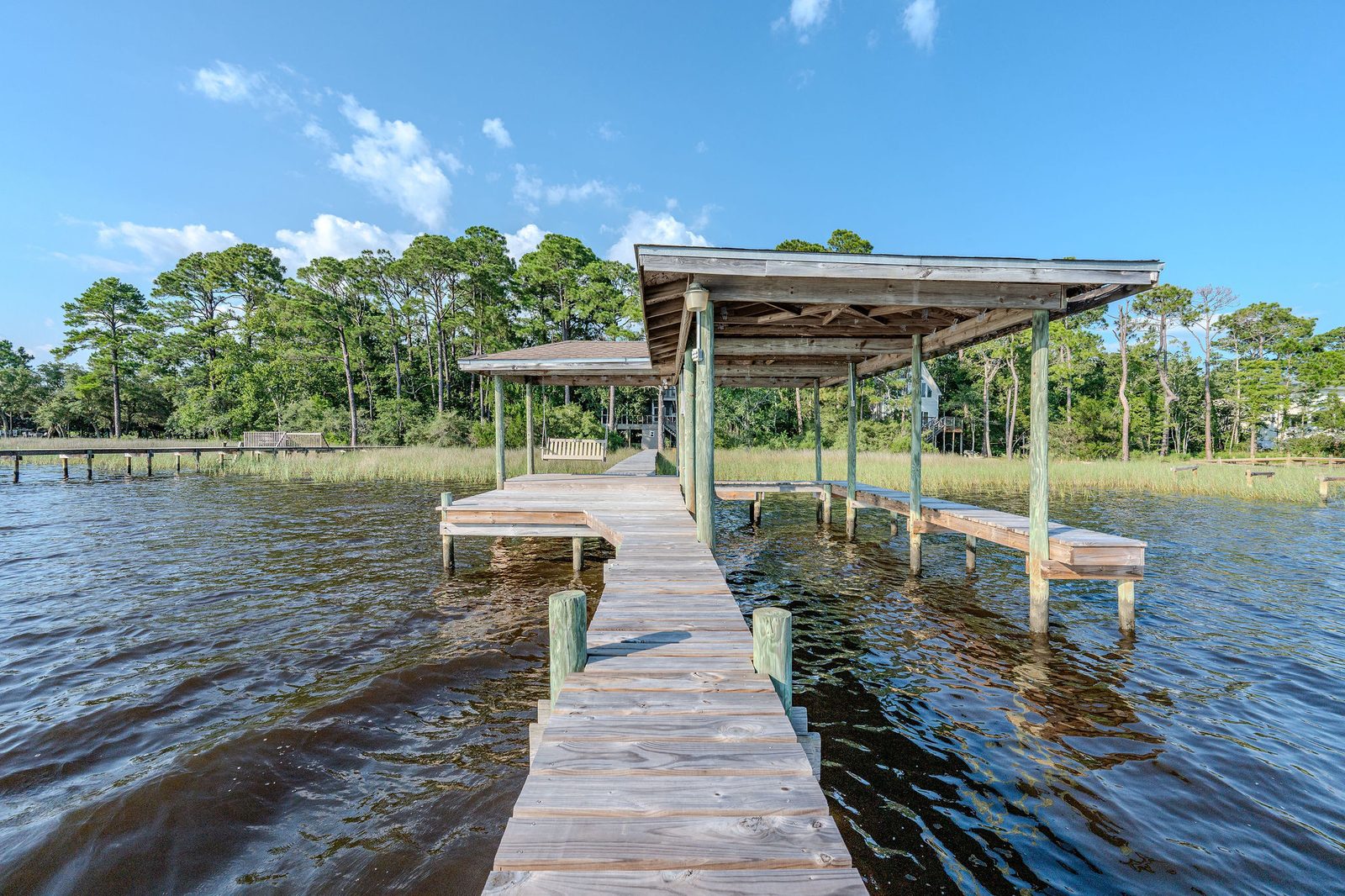 Looking back along a wooden pier toward shore: a covered boat slip with side catwalks on the right, a covered deck with a hanging swing on the left, and the house in the pines beyond the marsh.