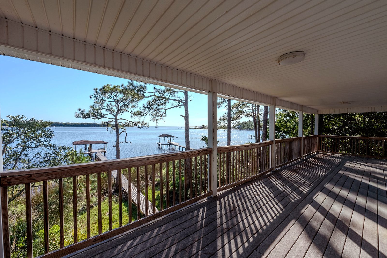 A long covered balcony with a wood rail and white posts, looking over pines and marsh to the boardwalk, the covered dock, and open bay water.