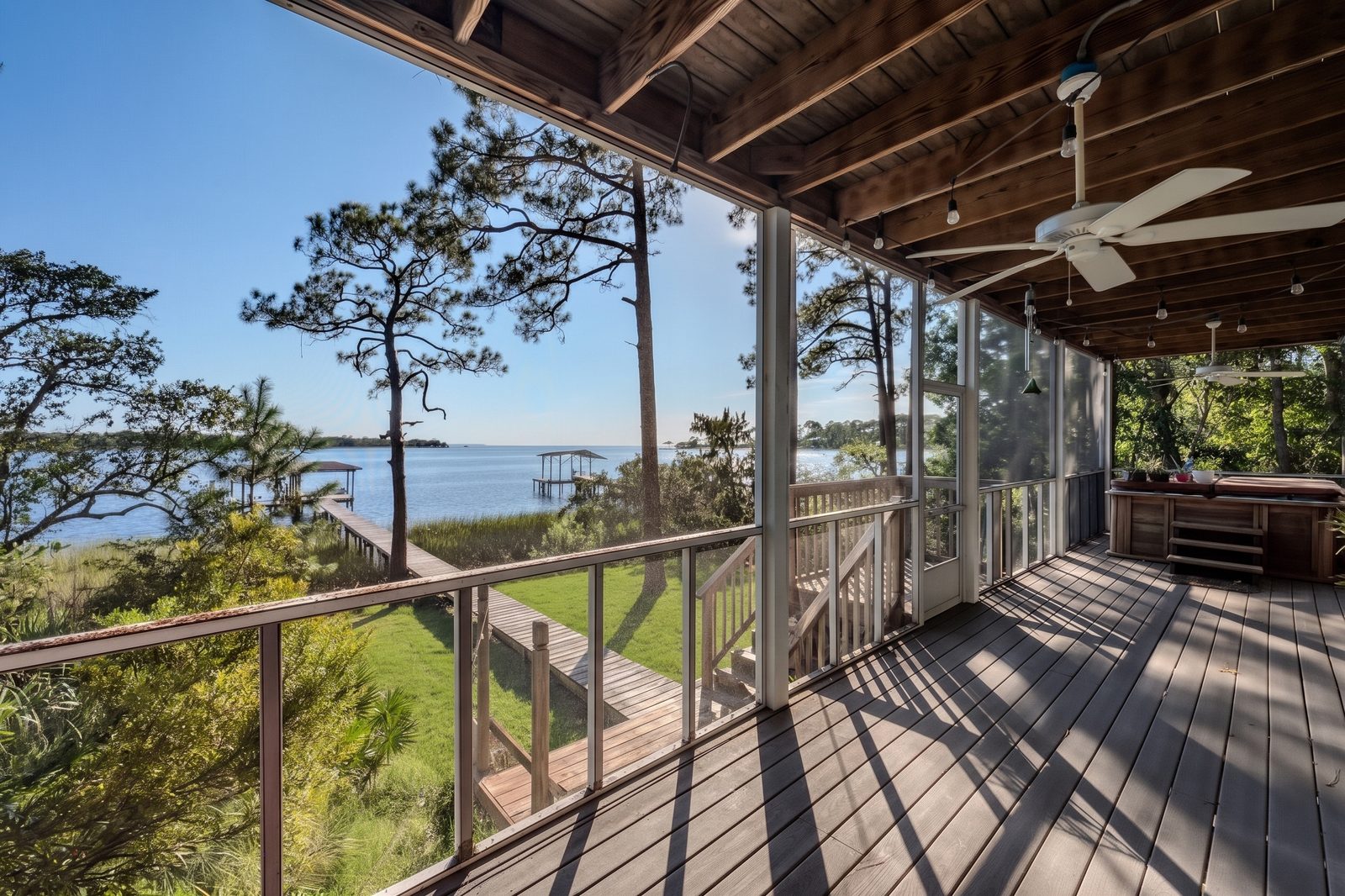 A screened porch with ceiling fans and string lights, looking over the lawn and the boardwalk to the covered dock and the bay, with a covered hot tub at the far end.