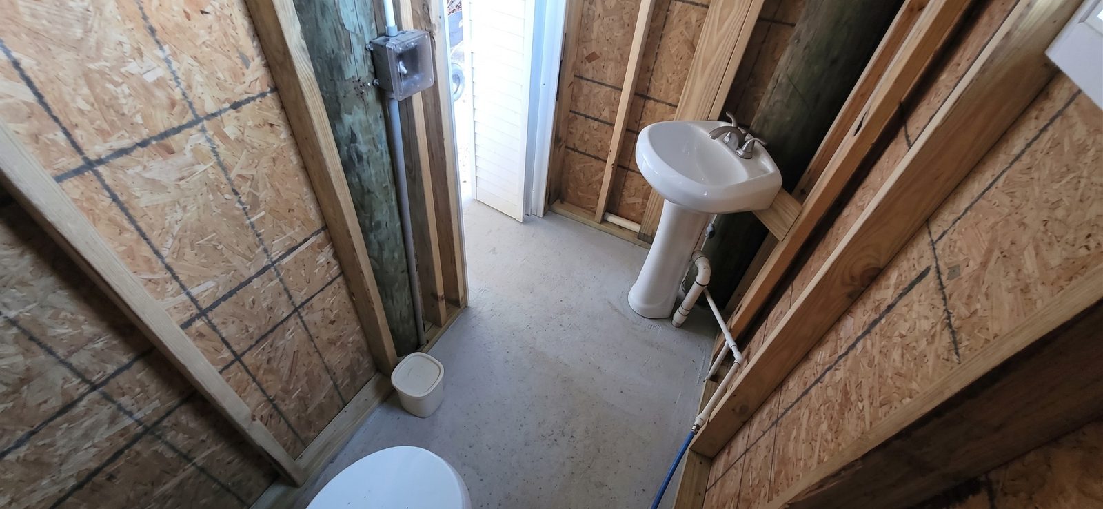 A small half bath with a concrete floor, a pedestal sink, and a toilet, its walls open wood framing and sheathing around a wood piling, with a door standing open to daylight.
