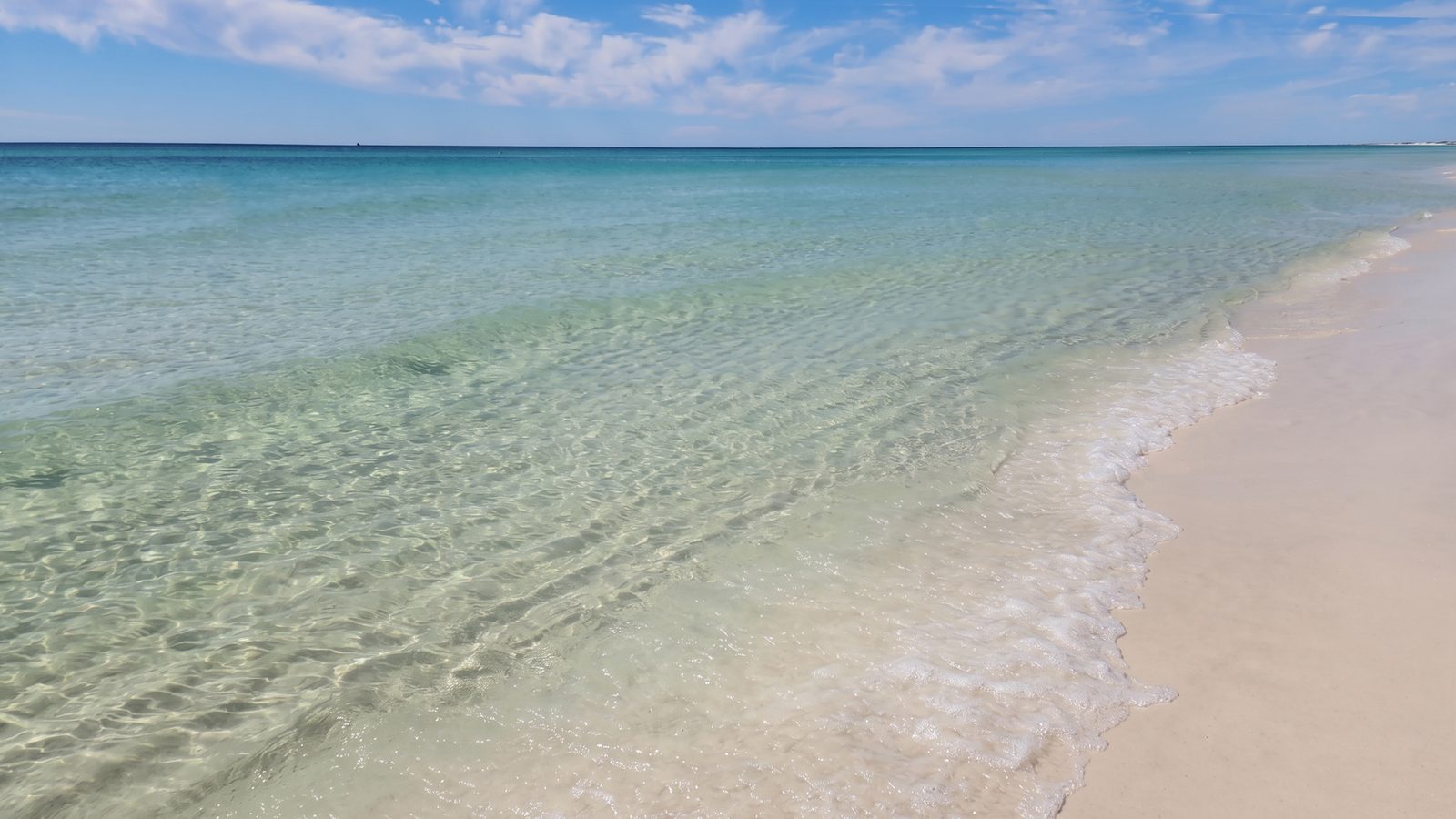 Clear, shallow turquoise water washing onto white sand at a beach, under a blue sky with light clouds.