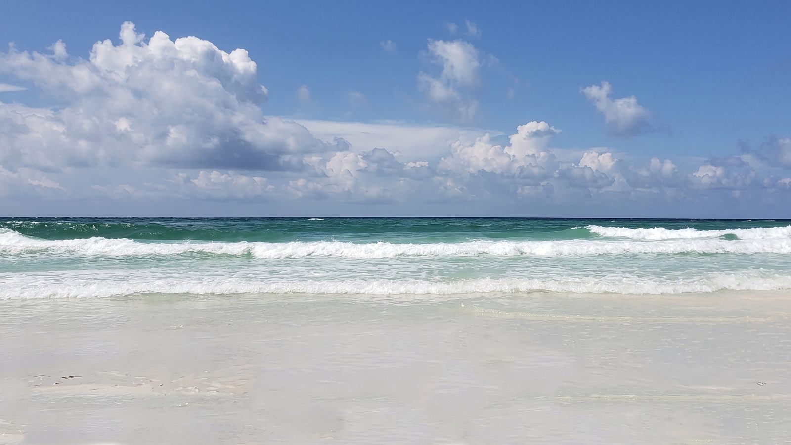Waves breaking onto a white-sand beach, green water out to the horizon, and tall white clouds in a blue sky.