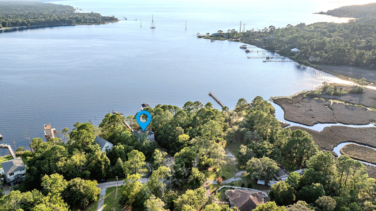 High aerial over East Bay: a blue location pin marks a house in the pines at the shoreline, with its boardwalk and covered dock at the water, open bay water out to a far wooded shore, and marsh and tidal creeks at right.