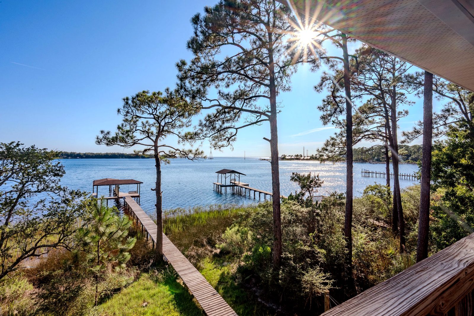 View from a covered balcony through tall pines: a boardwalk runs through marsh grass to the covered dock, with open bay water beyond.